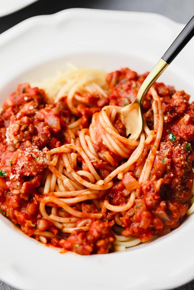 gold fork twirling tomato sauce-covered spaghetti noodles in a white bowl.
