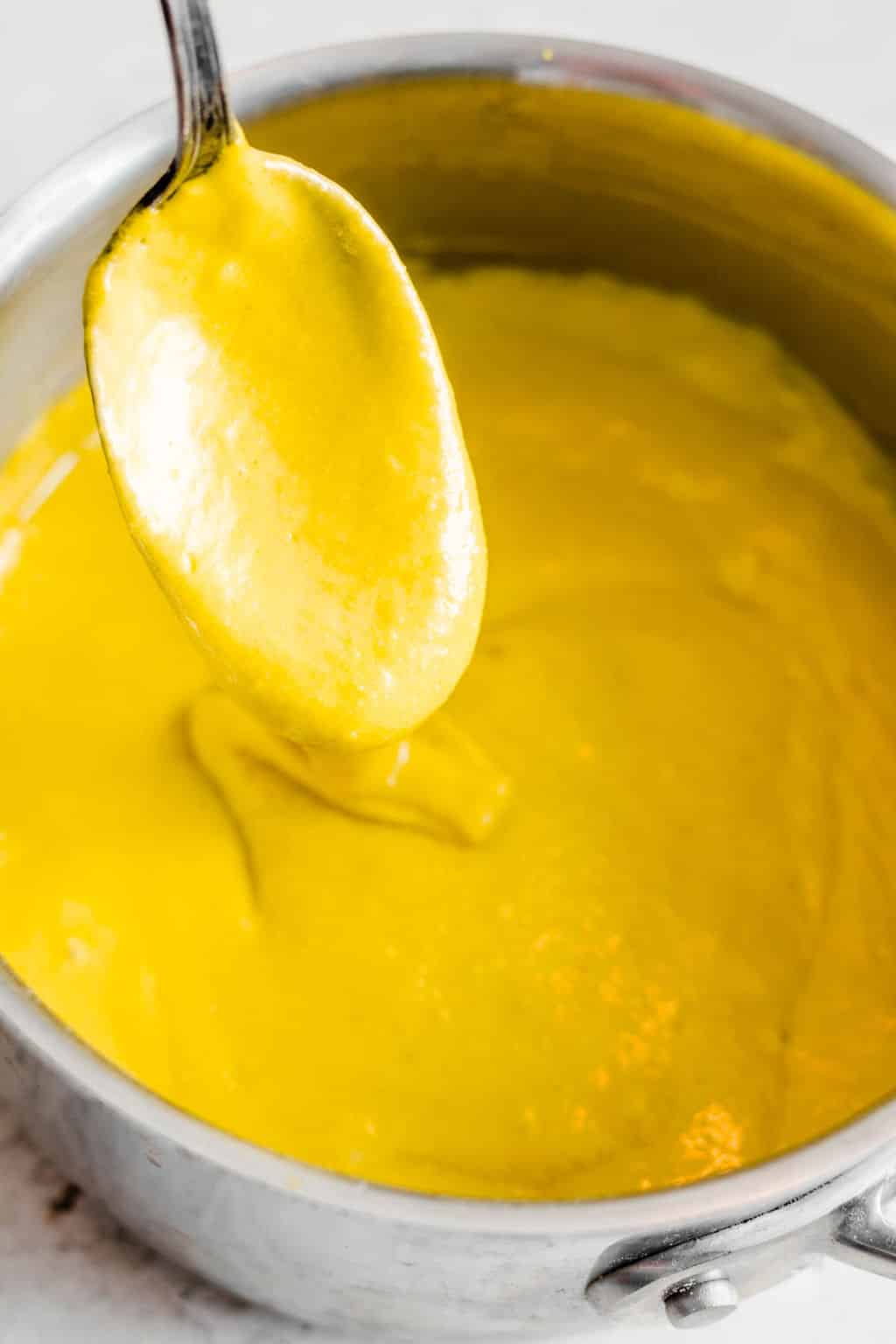 close up on a spoon taking a scoop of yellow cheese sauce out of a metal pot.
