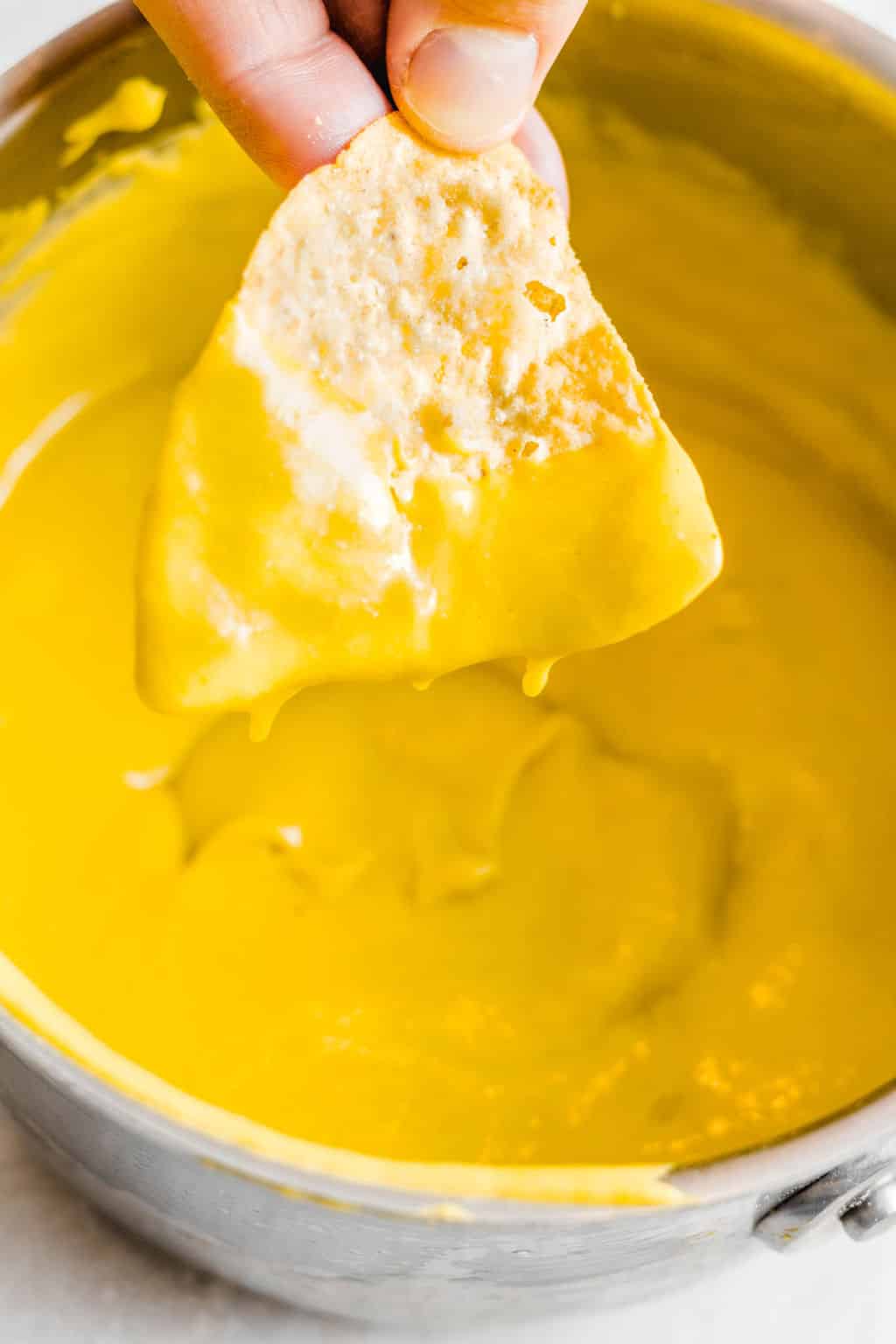 womans hand dipping a nacho chip into a pot filled with yellow cheese sauce.