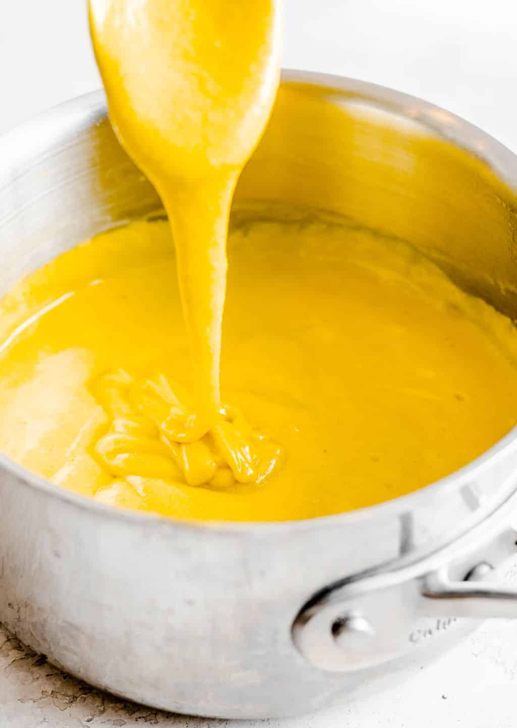 a stream of yellow cheese sauce dripping from a spoon back into a metal pot.
