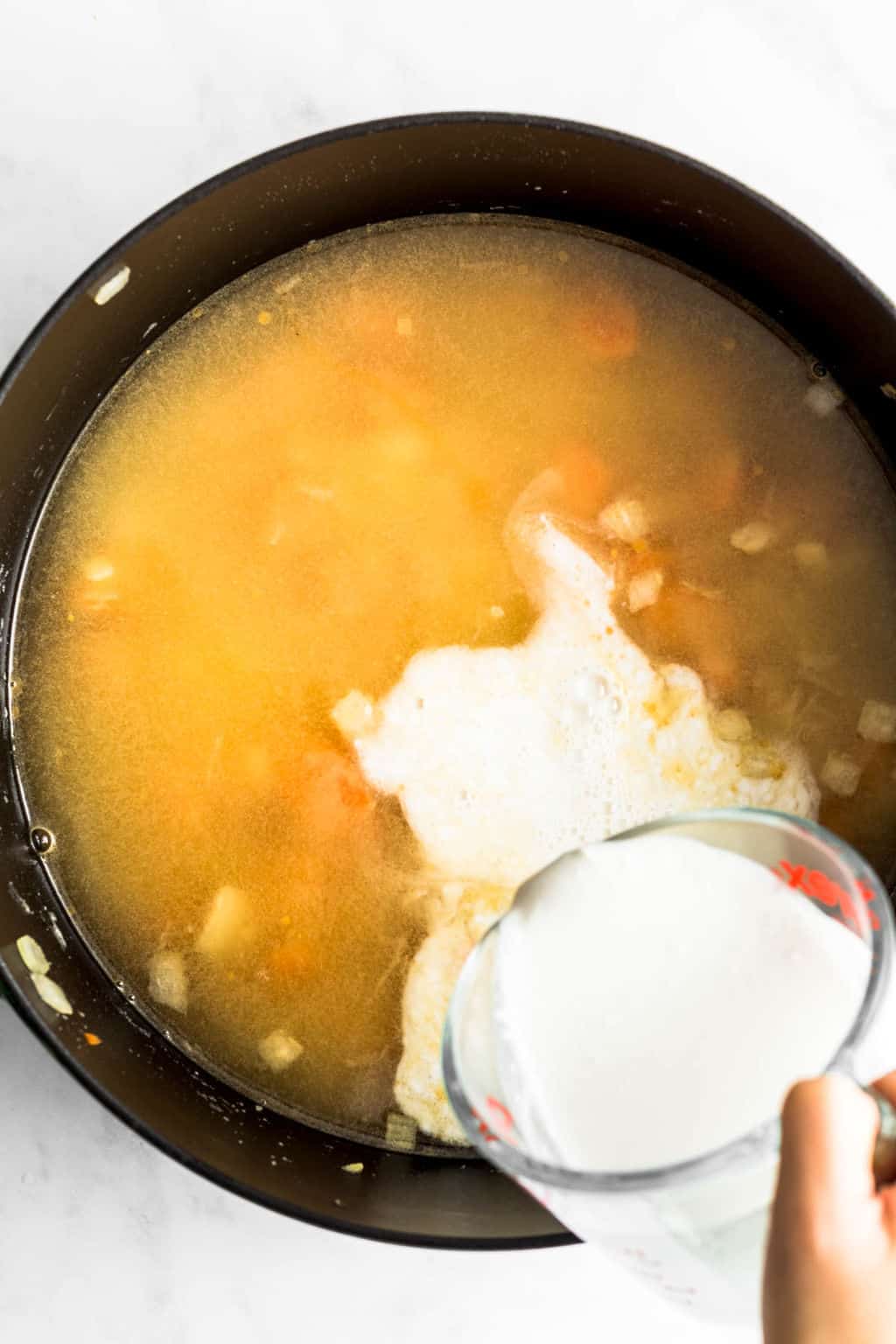 womans hand pouring white cream into a black pot filled with broth.