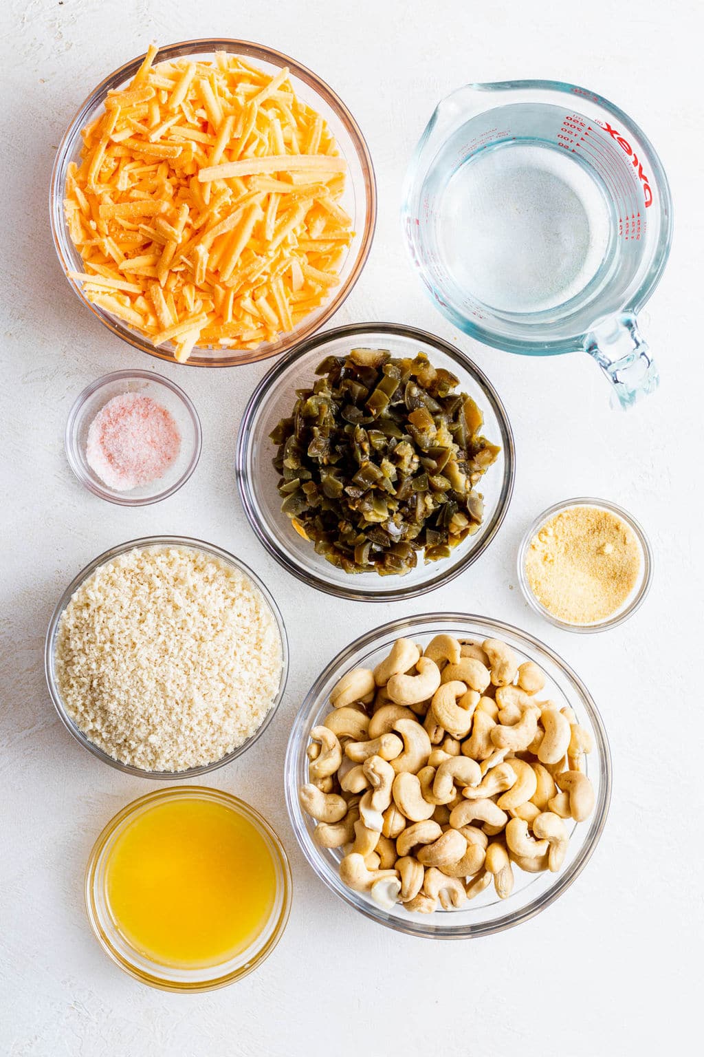 individual glass bowls holding vegan cheddar cheese, jalapeños, breadcrumbs, cashews, and melted butter.