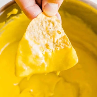 womans hand dipping a nacho chip into a pot filled with yellow cheese sauce.