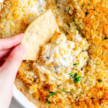 womans hand dipping a tortilla chip into a baked breadcrumb-covered white dip.