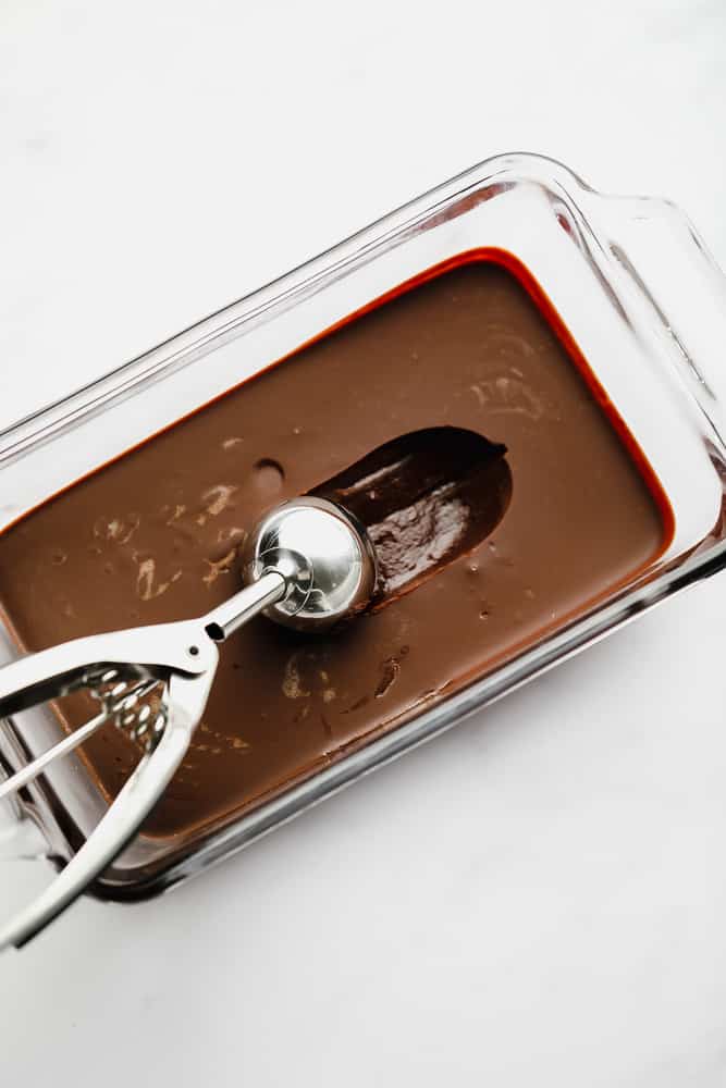 a metal ice cream scoop scooping out a ball of chocolate from a chocolate mixture in a glass dish.