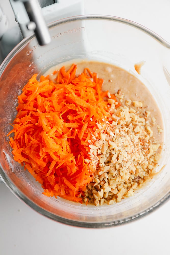 shredded carrots and walnuts being added to a clear bowl with batter