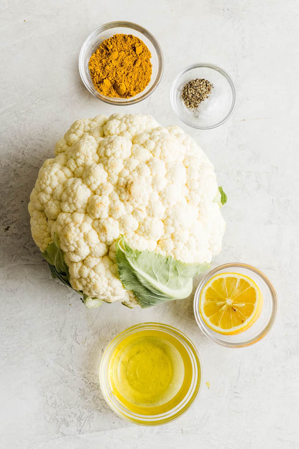a head of cauliflower next to small glass bowls filled with oil, lemon, pepper, and yellow curry powder.