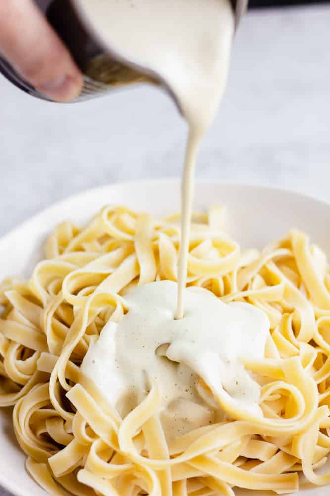 white sauce being poured over cooked noodles on white plate