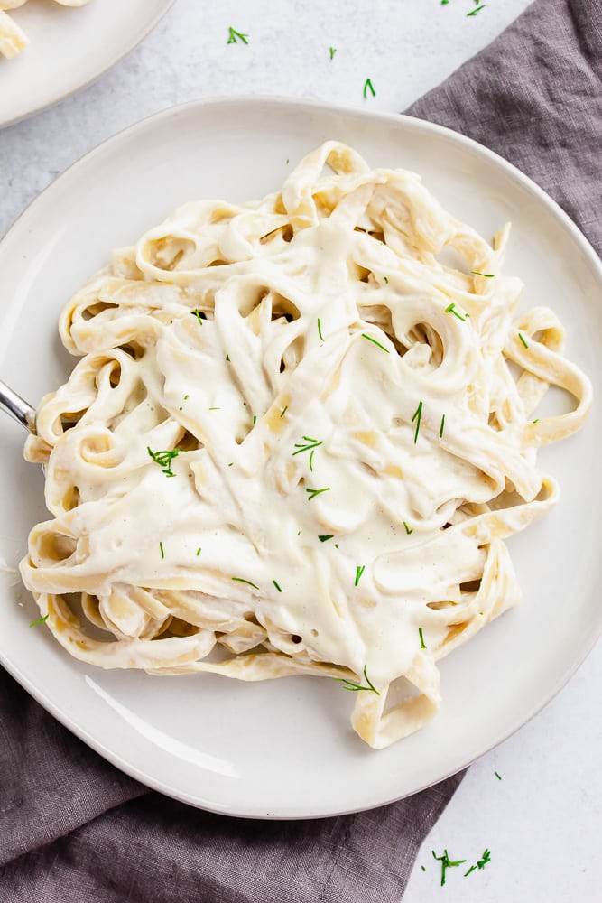 white plate with alfredo fettuccine, sprinkled with parsley and grey towel in background