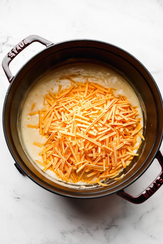 staub pot on white marble background, creamy sauce and shredded cheese added on top