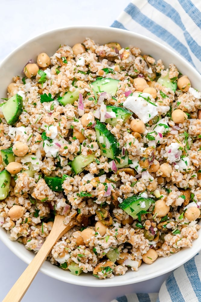 close up photo of a bowl of grain salad with wooden spoon and blue striped towel
