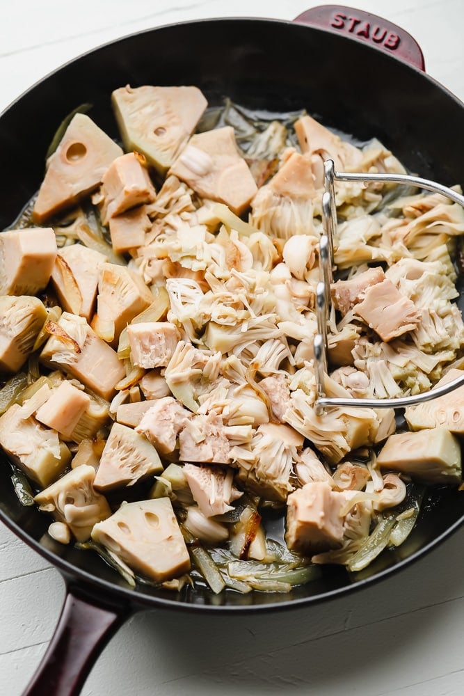 mashing jackfruit in a black skillet with a potato masher.