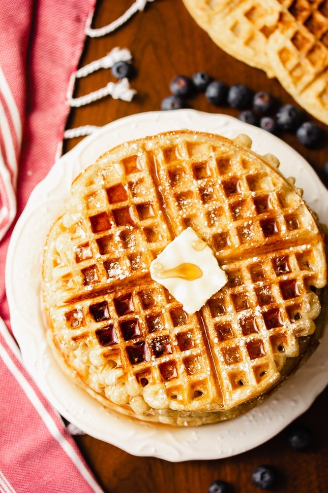 stack of waffles on a white plate with red towel and blueberries on wood backdrop