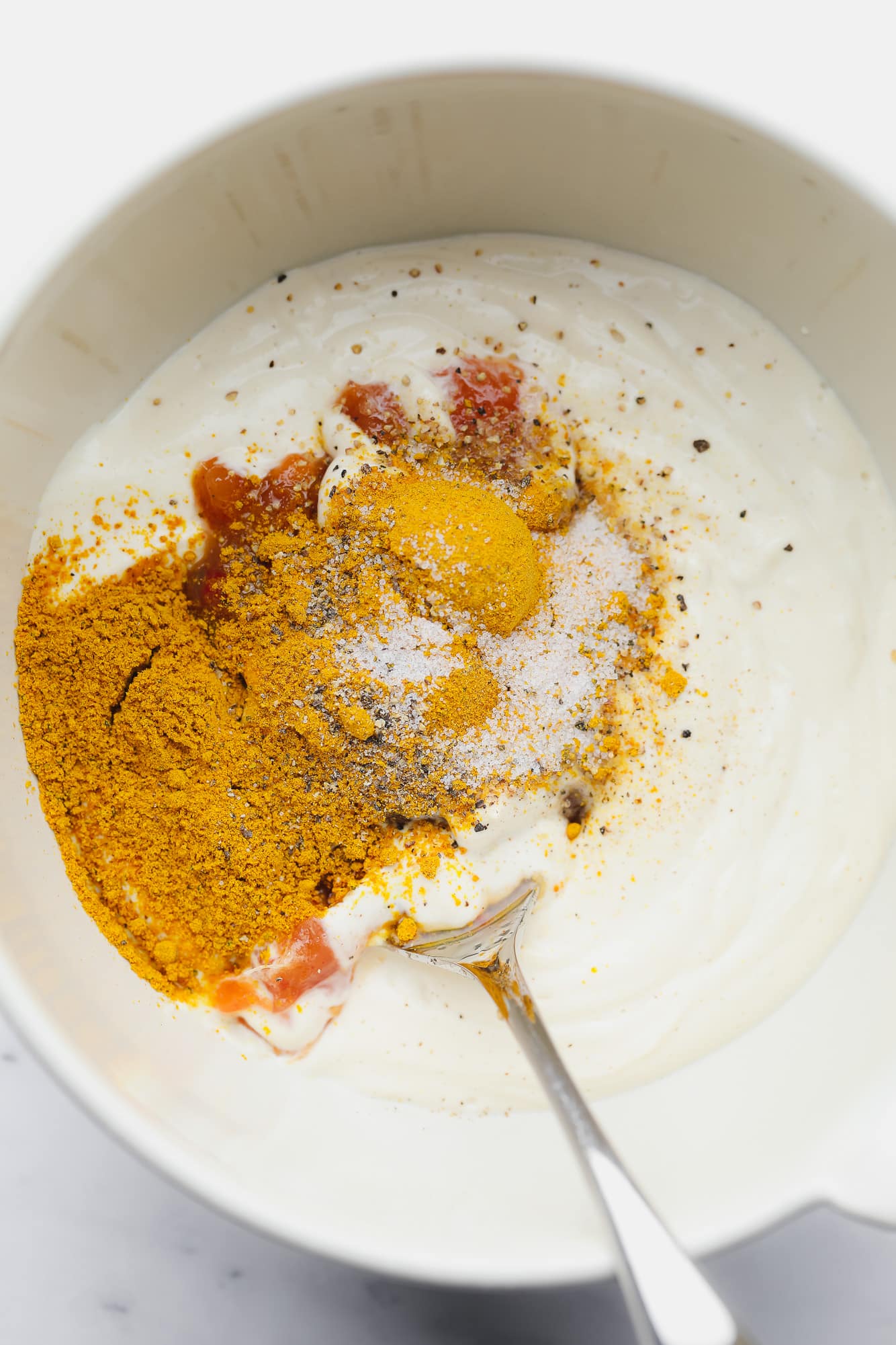 using a spoon to stir yellow and red spices into vegan mayonnaise in a white bowl.