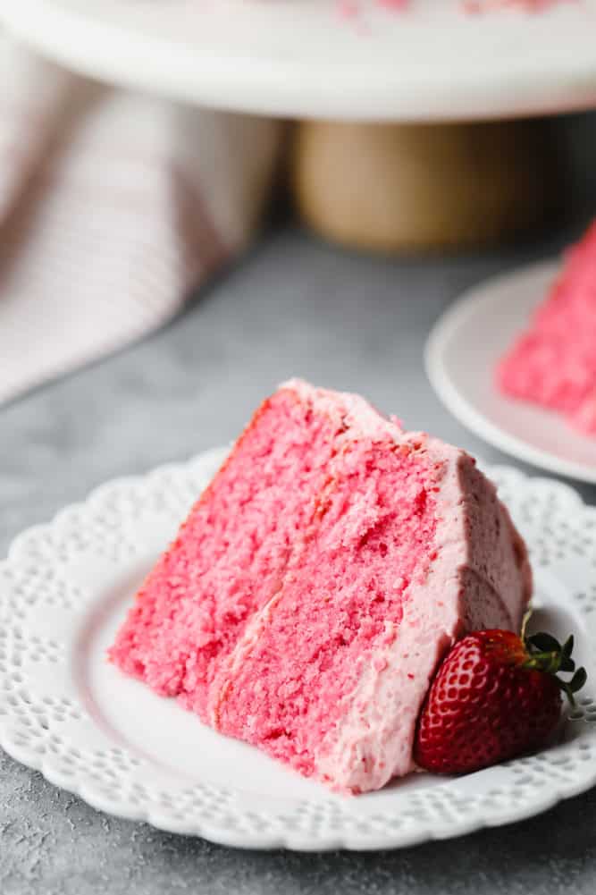 piece of pink cake on white plate, cake stand in background on grey board