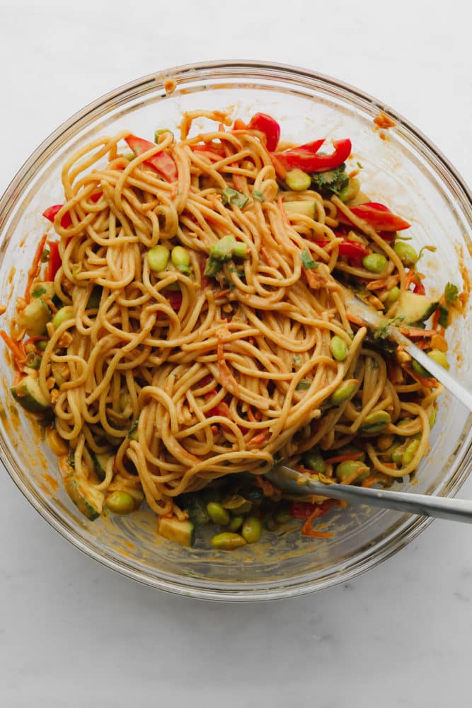 glass bowl with tongs stirring noodles with vegetables and peanut sauce