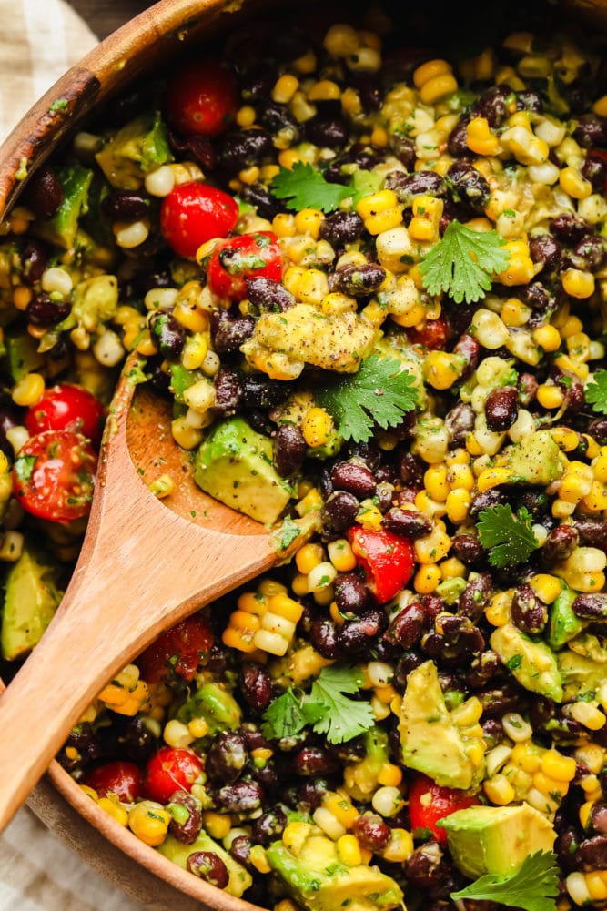 a wooden spoon scooping black bean salad out of a large bamboo bowl.