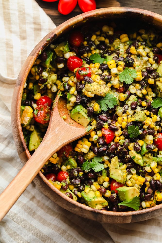 a wooden spoon scooping black bean salad out of a large bamboo bowl.