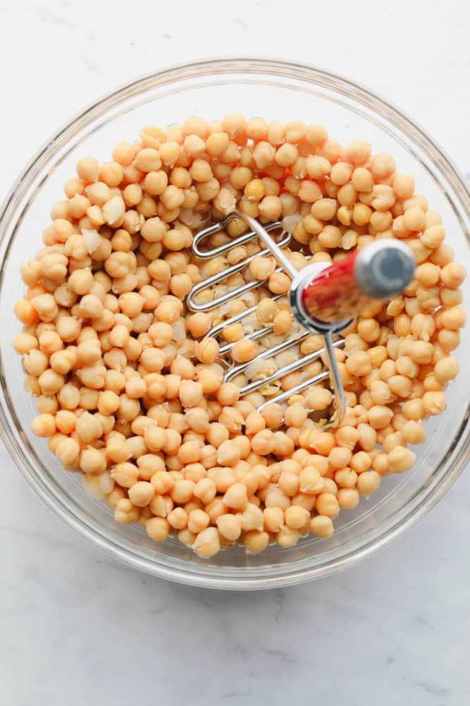 chickpeas being mashed in a bowl with potato masher