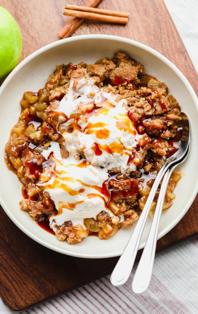 apple crisp with vegan salted caramel sauce and ice cream, two spoons in bowl and on a wood board.