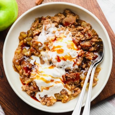 scoops of vegan apple crumble with vanilla ice cream on top in a white bowl with 2 spoons.