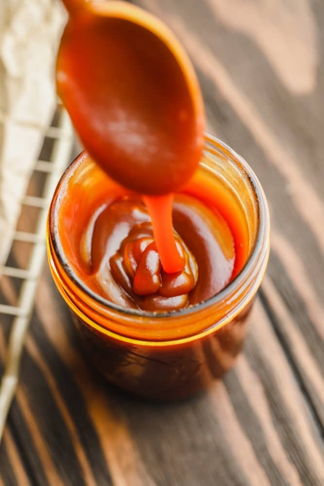caramel sauce in a glass jar being drizzled down with a spoon, wood backdrop.