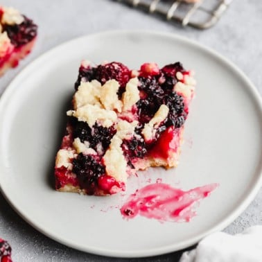 square image of a blackberry bar with shortbread crumble and a bite taken out of it on grey plate