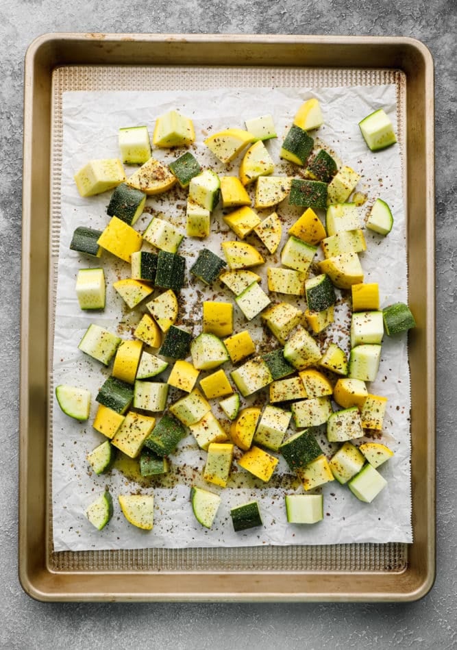 parchment lined gold pan with uncooked chunks of yellow squash and zucchini with seasonings on grey background.