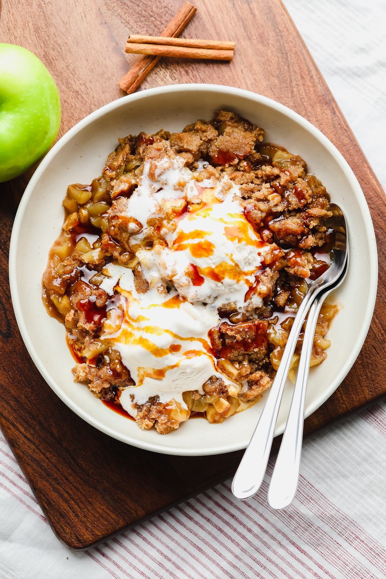 scoops of vegan apple crumble with vanilla ice cream on top in a white bowl with 2 spoons.