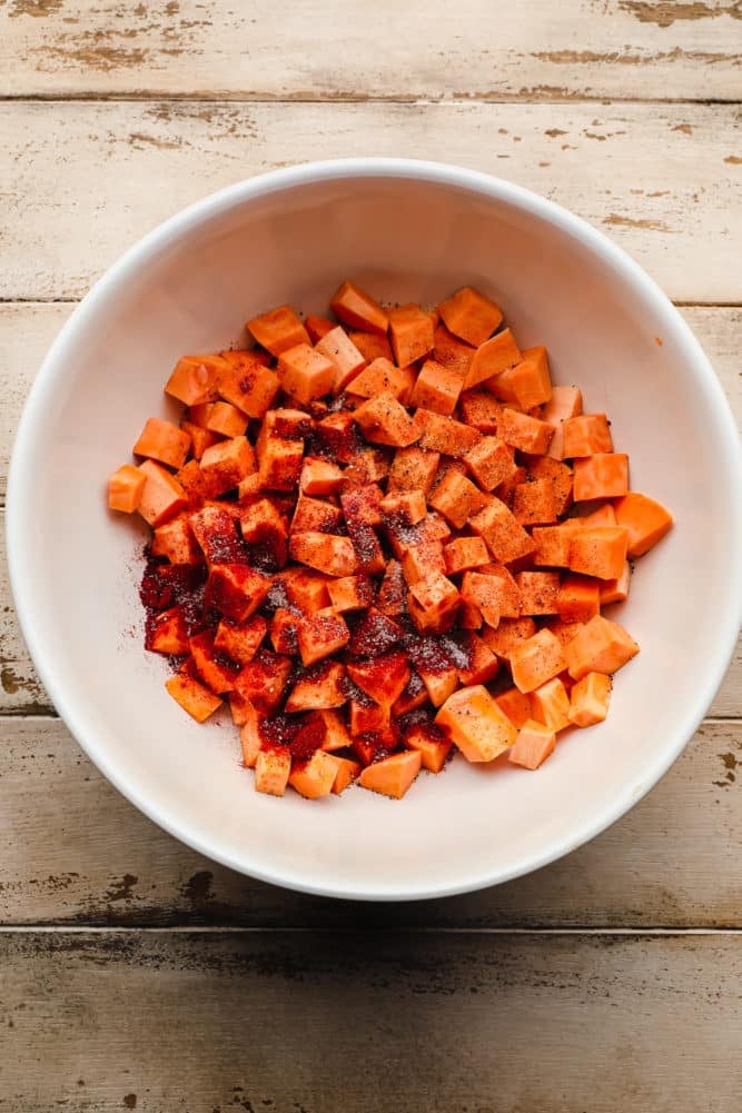 white bowl full of raw chopped and peeled sweet potatoes and spices on wood background.