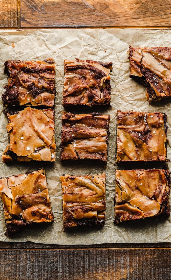 lots of brownies on a wooden background with parchment paper and caramel on top of them.