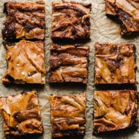 9 vegan salted caramel brownies on parchment paper, one with a bite taken out of it.