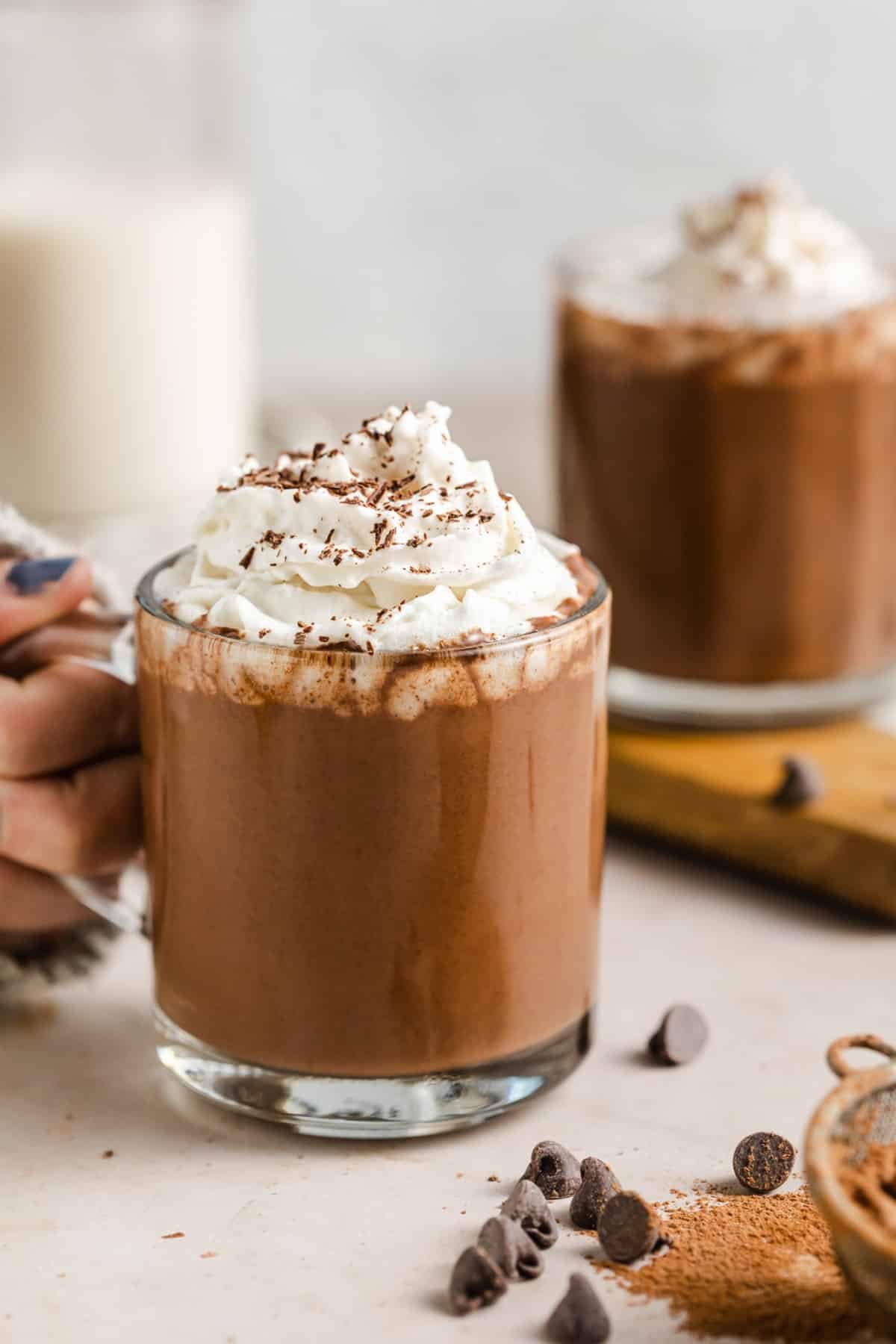 a woman holding onto a glass mug filled with vegan hot chocolate topped with whipped cream.