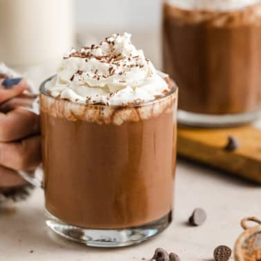 a woman holding onto a glass mug filled with vegan hot chocolate topped with whipped cream.