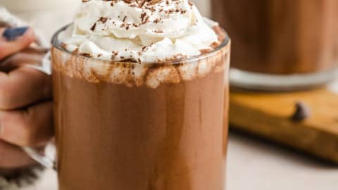 a woman holding onto a glass mug filled with vegan hot chocolate topped with whipped cream.