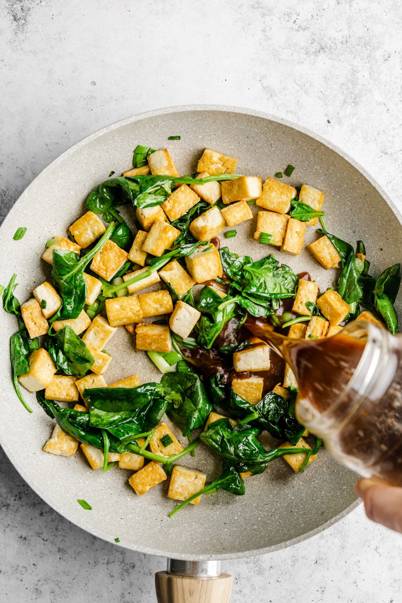 Pouring stir fry sauce in a grey skillet with tofu and spinach.