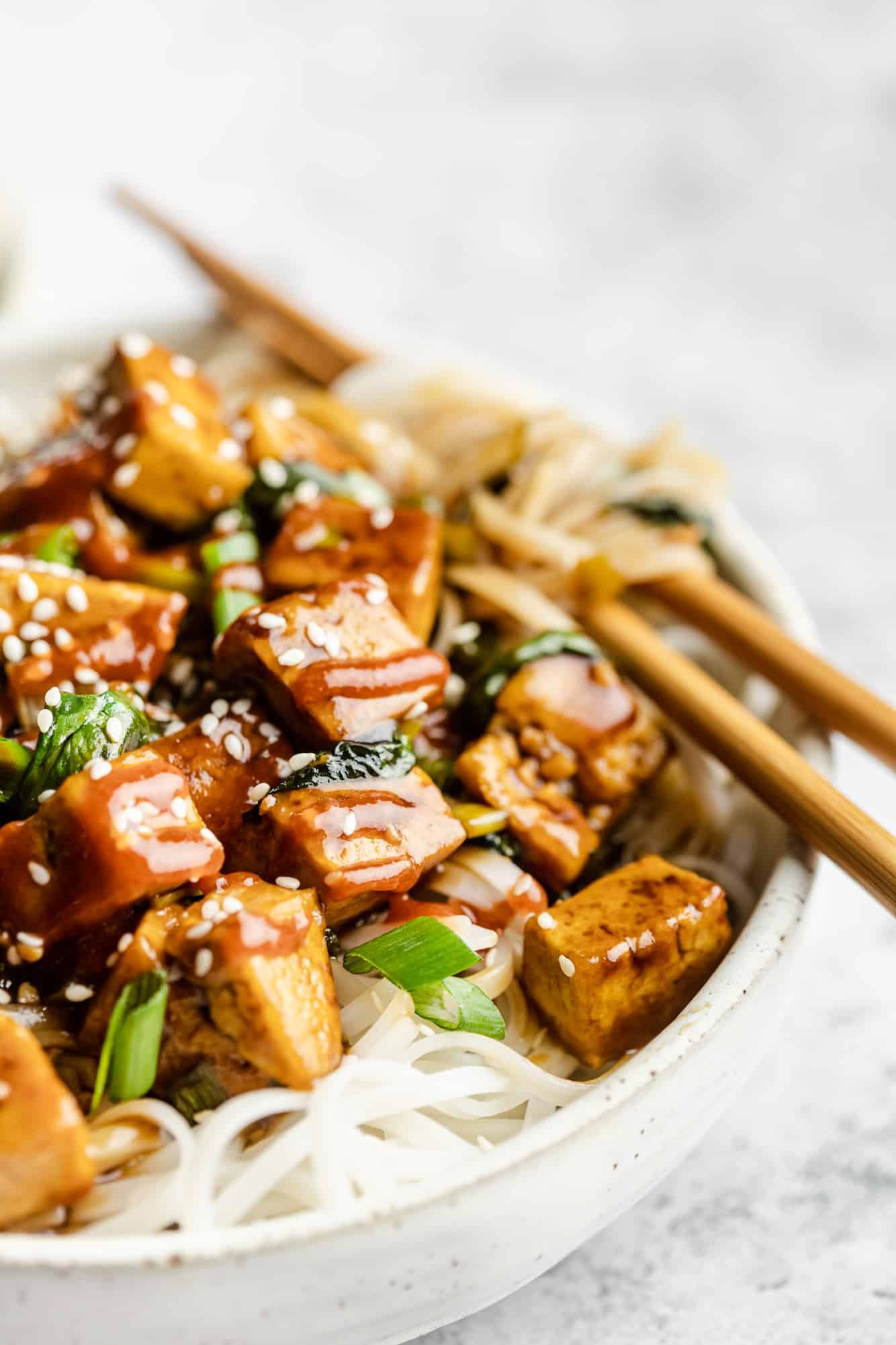 close up on tofu stir fry over rice noodles in a bowl with chop sticks.