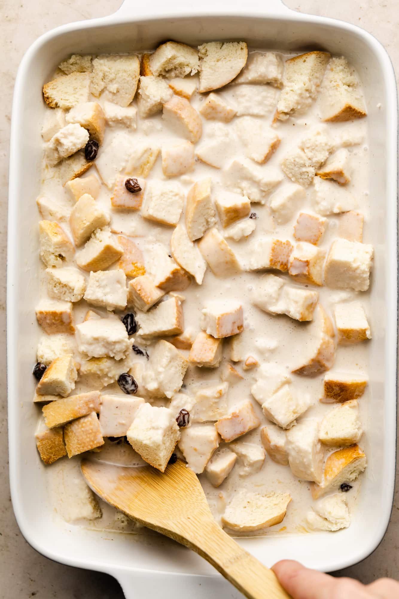 womans hand using a wooden spoon to stir bread cubes with white sauce in a white baking dish.