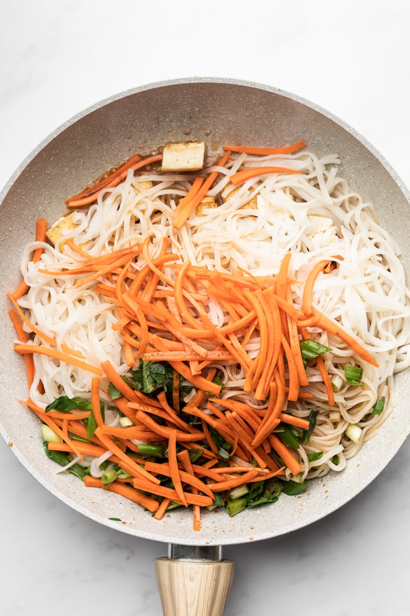 cooking rice noodles, carrots, and scallions together in a beige pan.