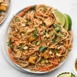 a plate of vegan pad thai topped with peanuts, lime wedges, and cilantro.
