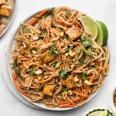 a plate of vegan pad thai topped with peanuts, lime wedges, and cilantro.