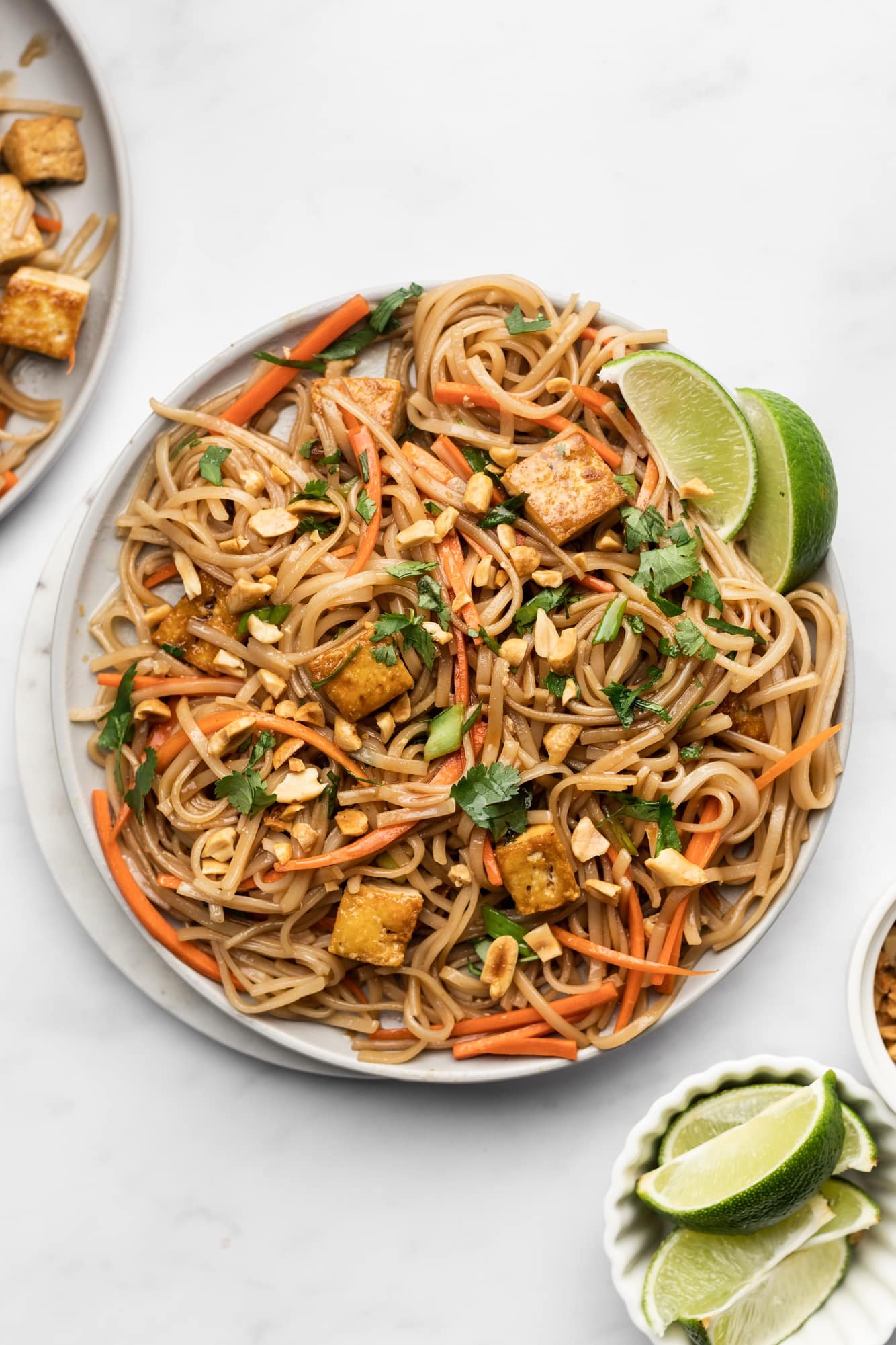 a plate of vegan pad thai topped with peanuts, lime wedges, and cilantro.