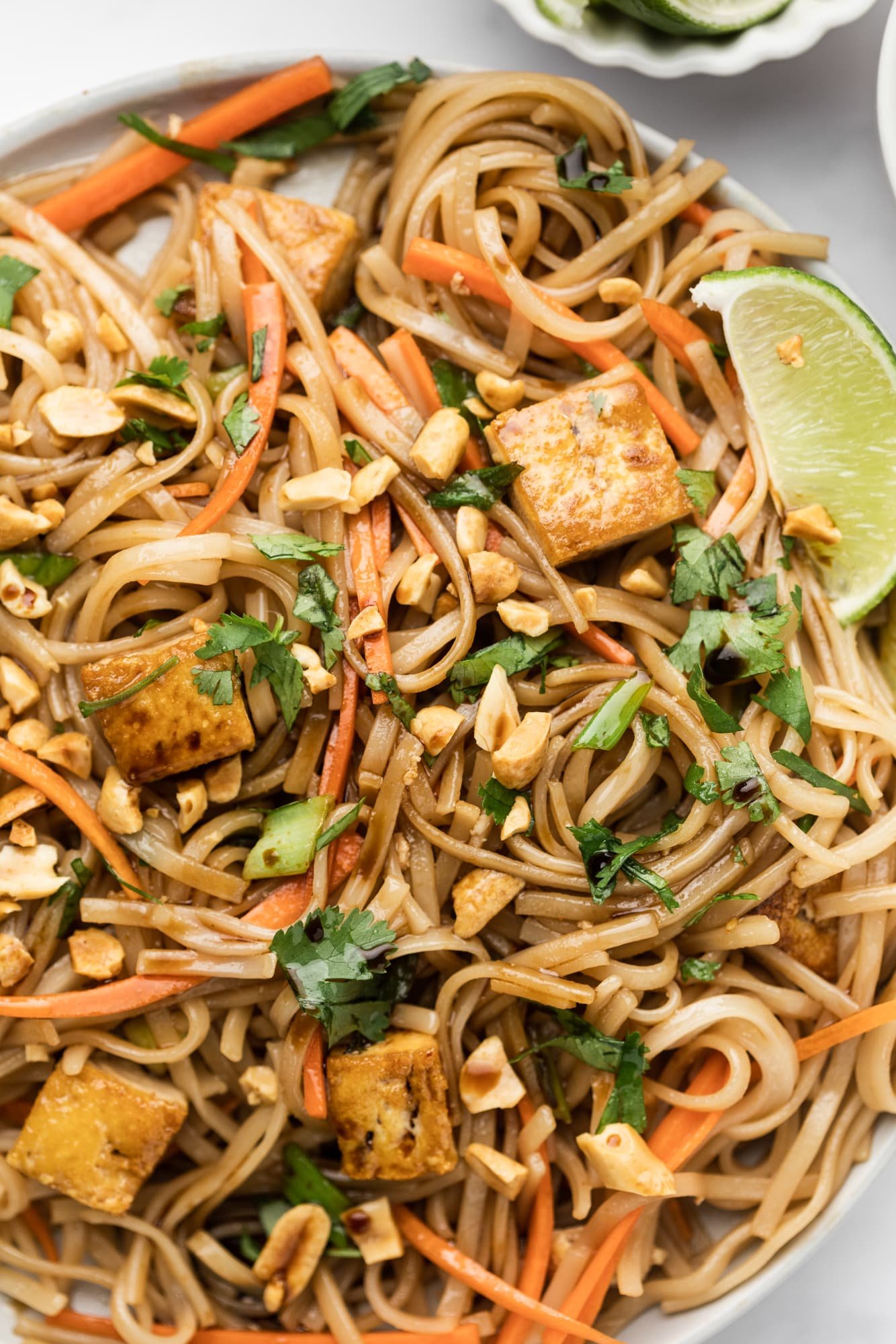 Close up on a plate of vegan pad thai topped with peanuts, lime wedges and cilantro.