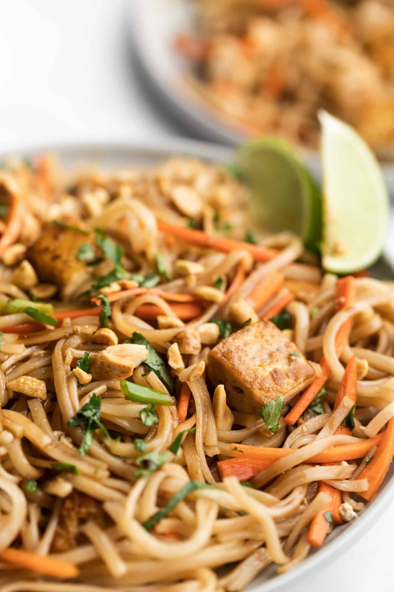 Close up on a plate of vegan pad thai topped with peanuts, lime wedges and cilantro.
