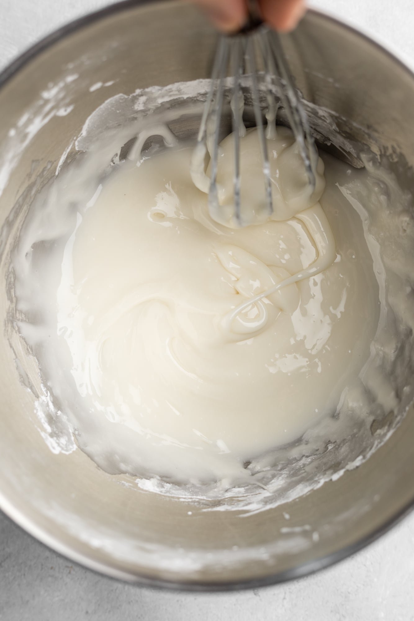 using a whisk to stir white icing in a large metal bowl.