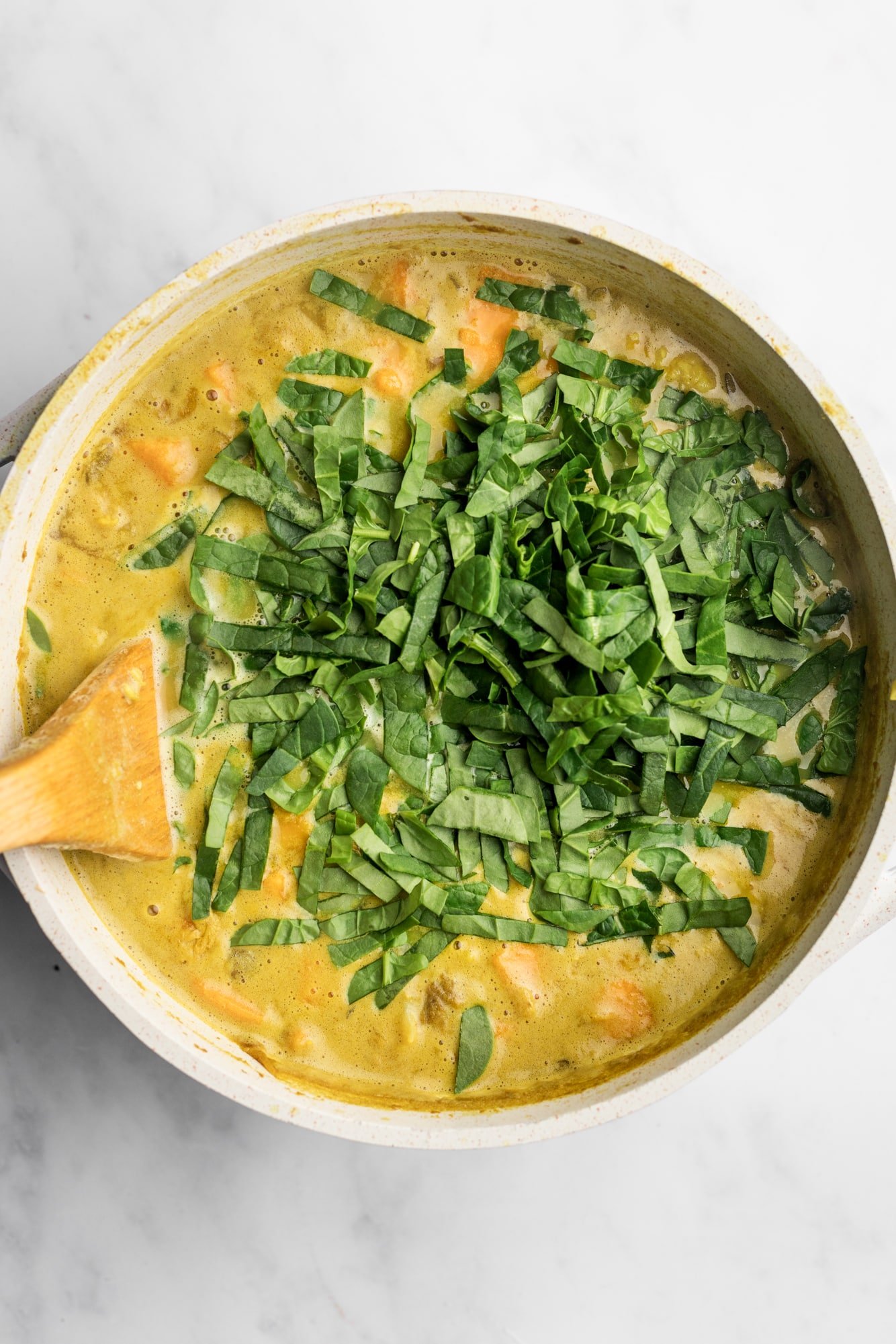 using a wooden spoon to stir spinach into a large pot of coconut curry soup.