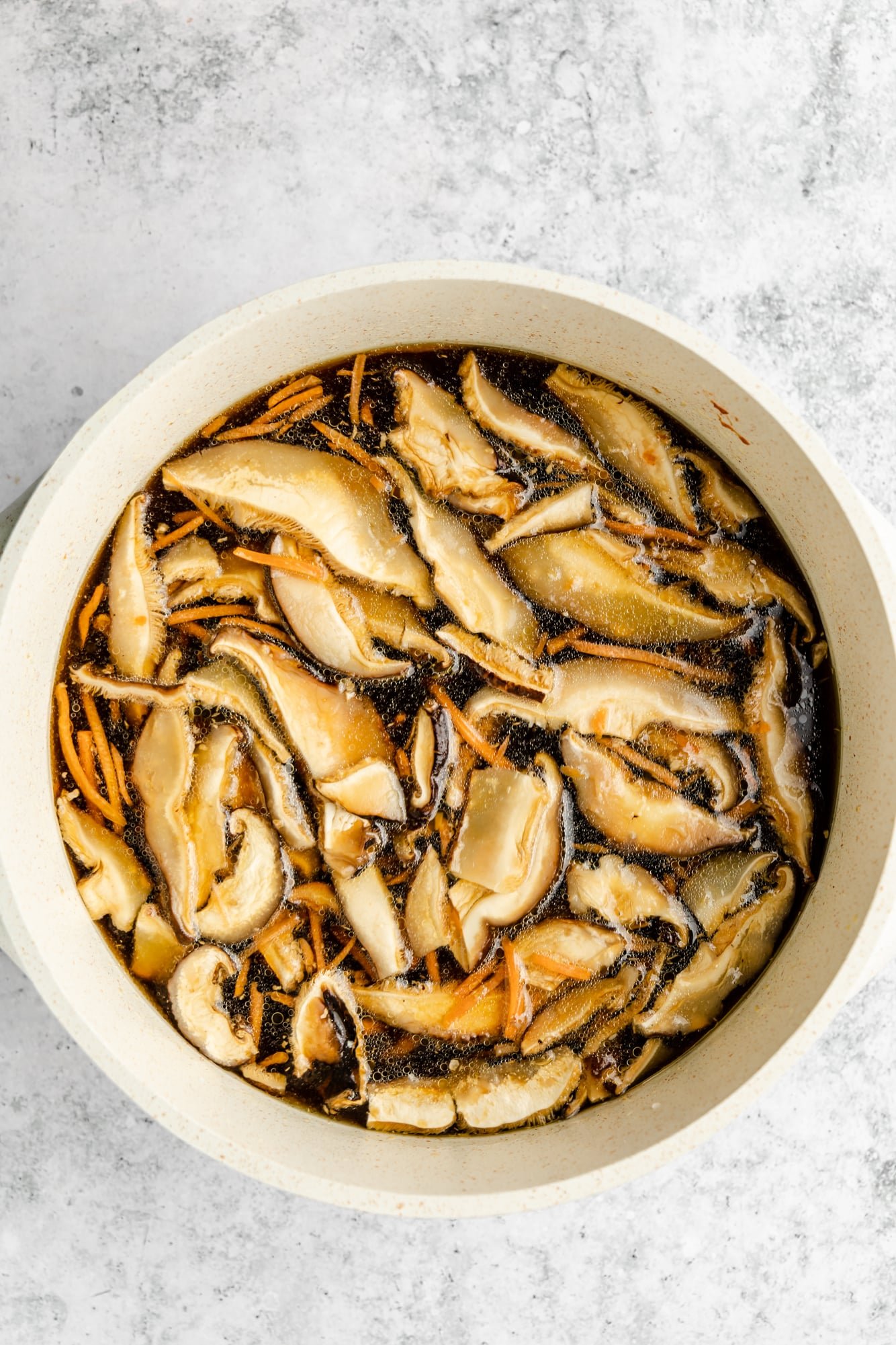 mushrooms and carrots simmering in a dark brown broth in a large white pot.