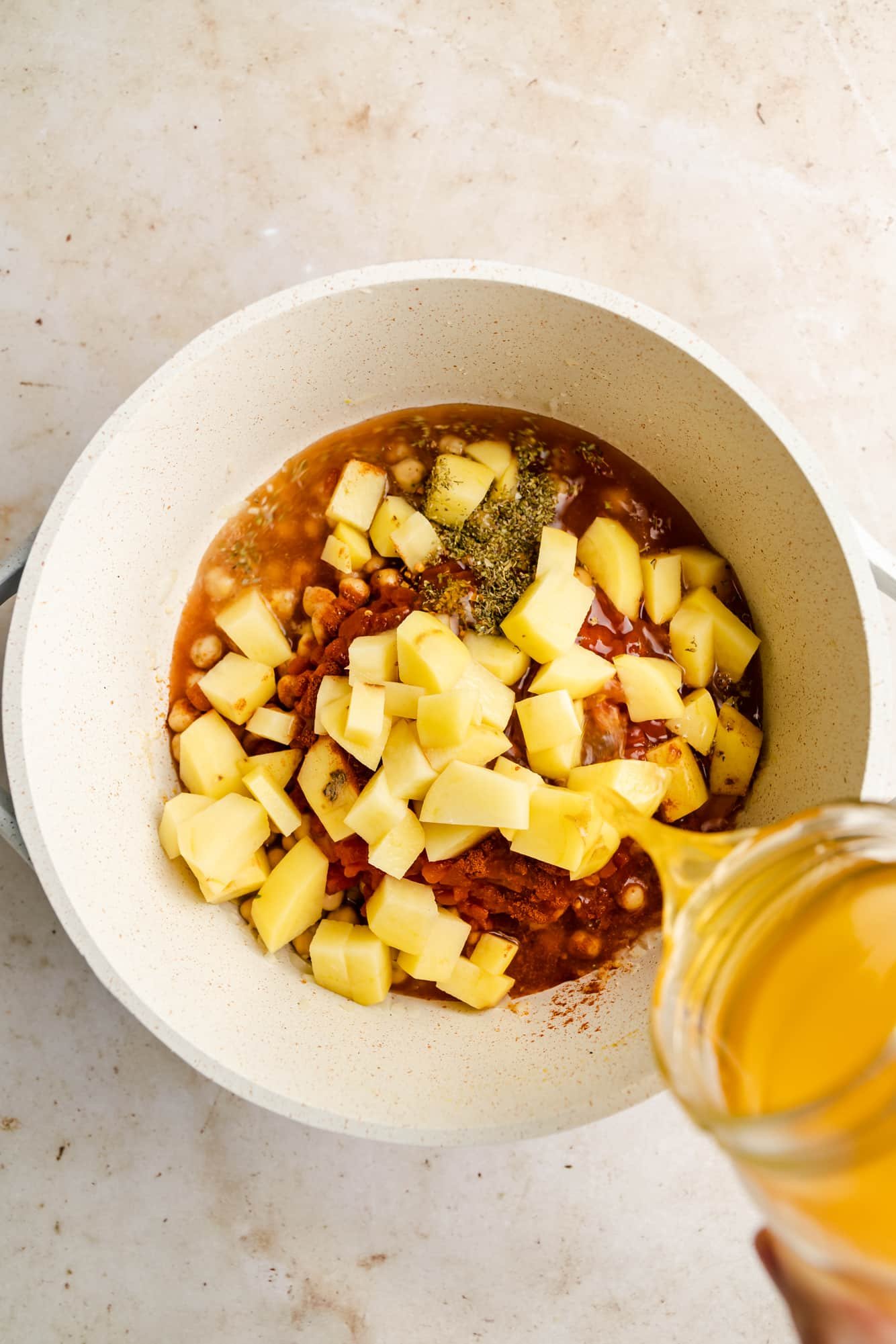 pouring broth into a white pot full of diced potatoes, herbs, tomatoes, and chickpeas.