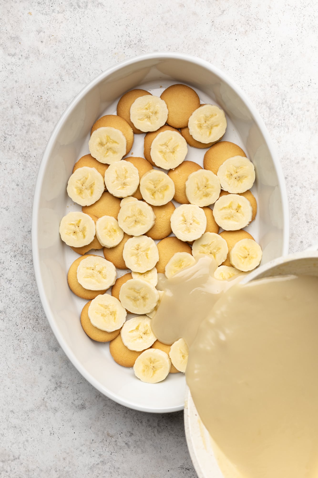 pouring vegan vanilla pudding over layers of vanilla cookies and banana slices in a large white dish.