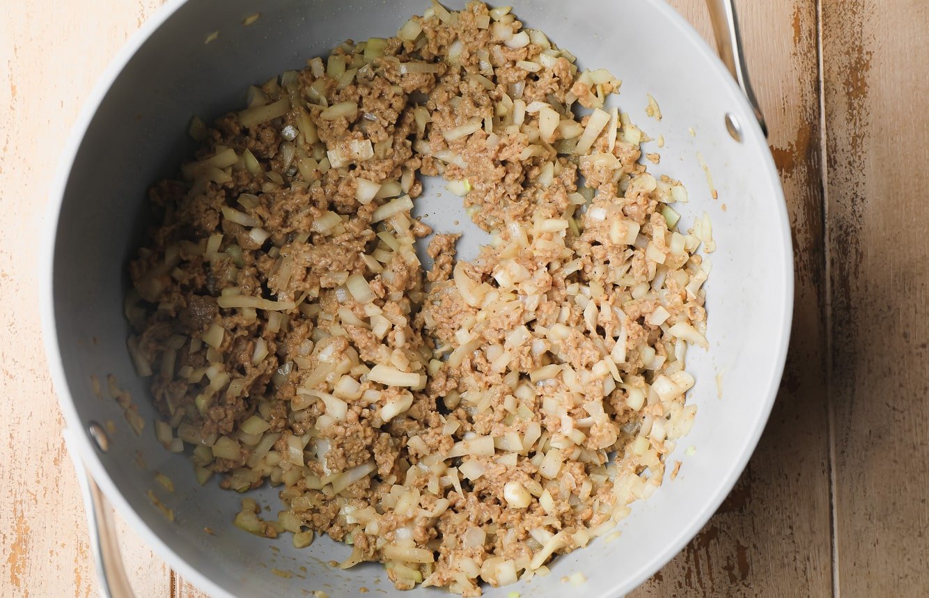 sauteeing onions and vegan sausage crumbles in a large grey pot.